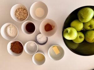 Overhead view of apple crisp ingredients including Granny Smith apples, oats, dark brown sugar, flour, cinnamon, butter, vanilla, lemon juice, and salt.