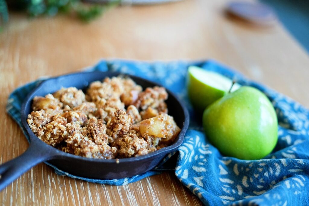 Cast iron skillet of apple crisp with a spoon resting inside and a serving of crisp plated next to it.
