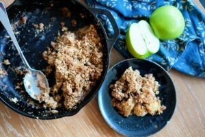 Overhead image of apple crisp in a mini cast iron skillet next to sliced green apples on a wooden table.