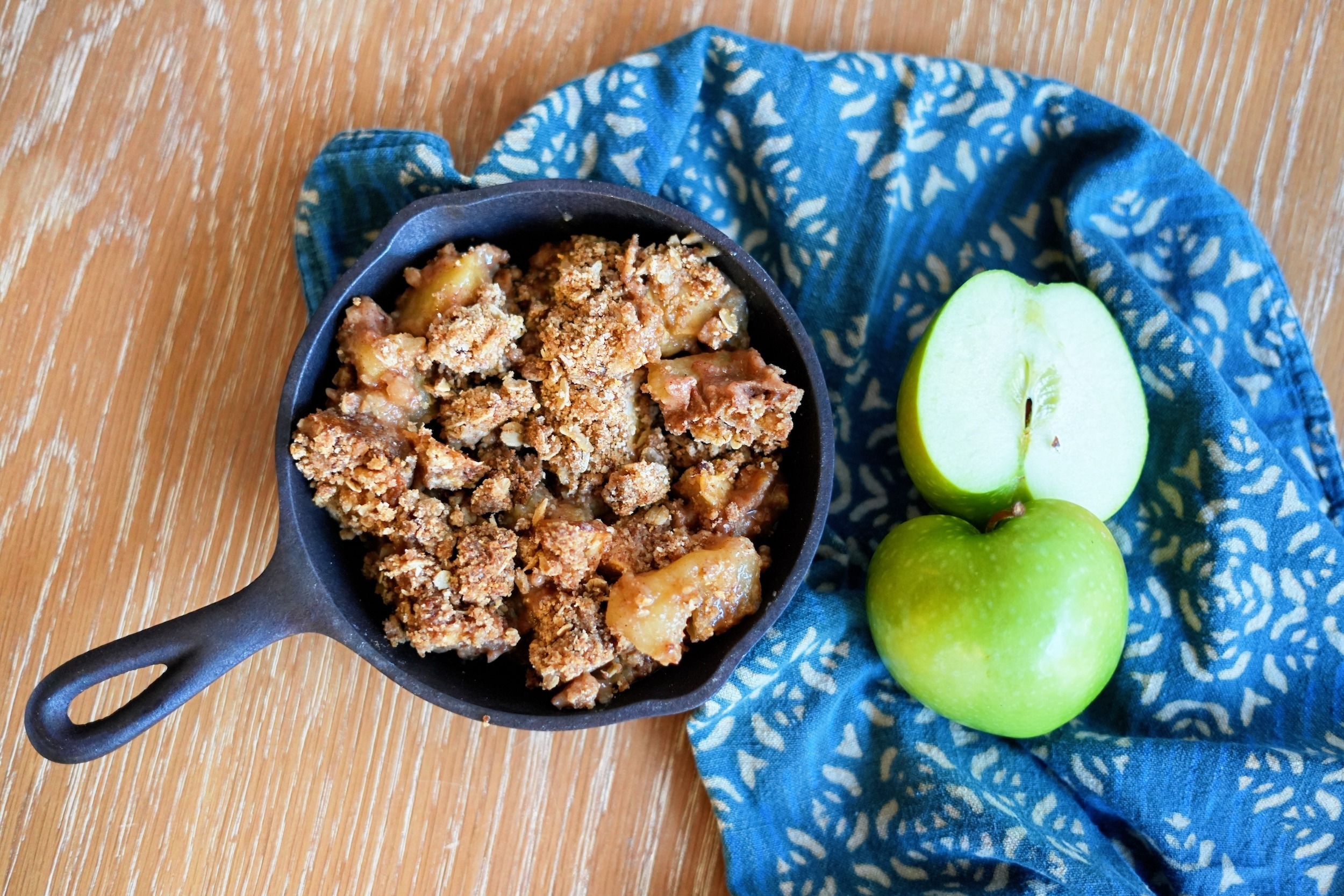 Small cast iron skillet filled with warm apple crisp, topped with oat crumble, styled with green apples and a blue cloth.