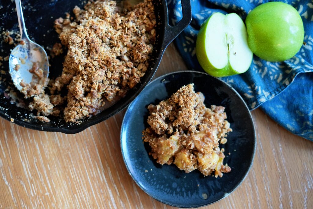 Cast iron skillet of apple crisp with a spoon resting inside and a serving of crisp plated next to it.
