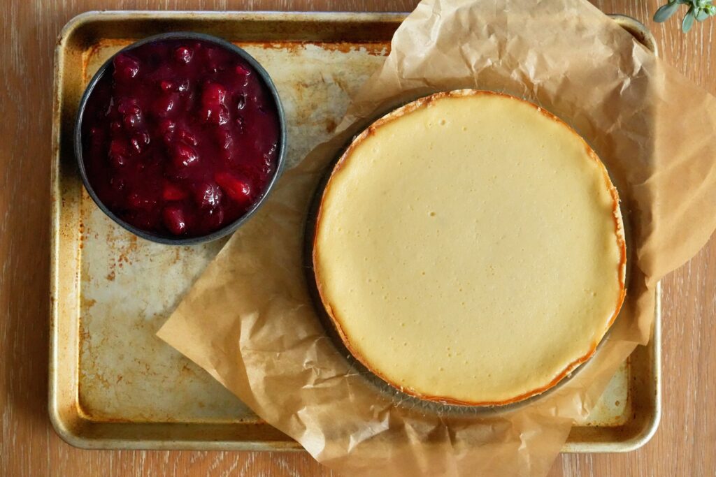 Plain baked cheesecake next to a bowl of guava cranberry sauce on parchment paper.