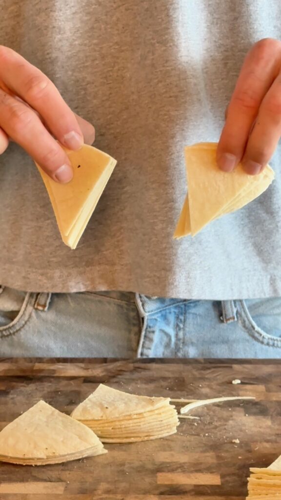 Slicing corn tortillas into triangles on a wooden cutting board to prepare homemade Cool Ranch chips.
