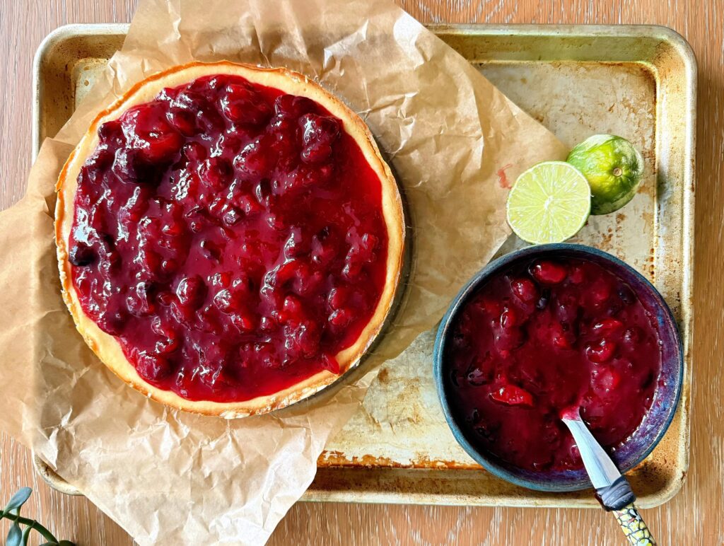 Overhead view of guava cranberry cheesecake with bright red fruit topping and lime on a sheet pan.