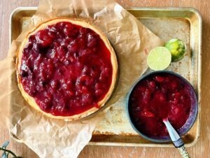 Overhead view of guava cranberry cheesecake with bright red fruit topping and lime on a sheet pan.
