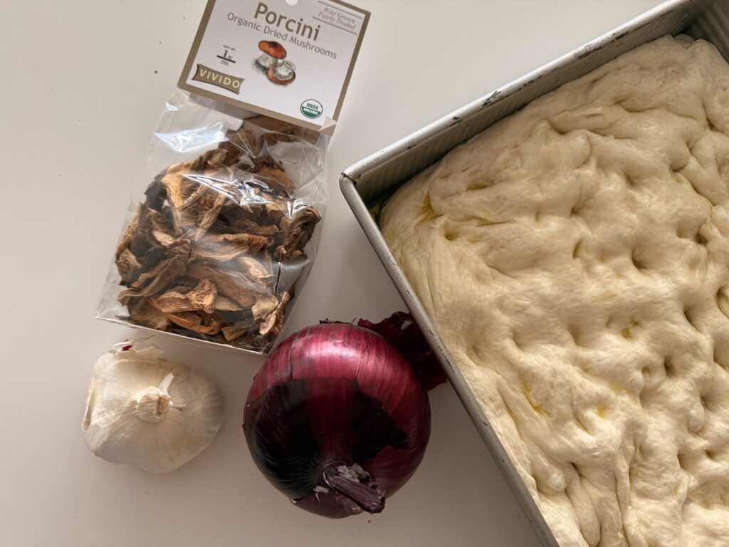 Ingredients for porcini focaccia including dried porcini mushrooms, garlic, red onion, and risen dough in a pan.