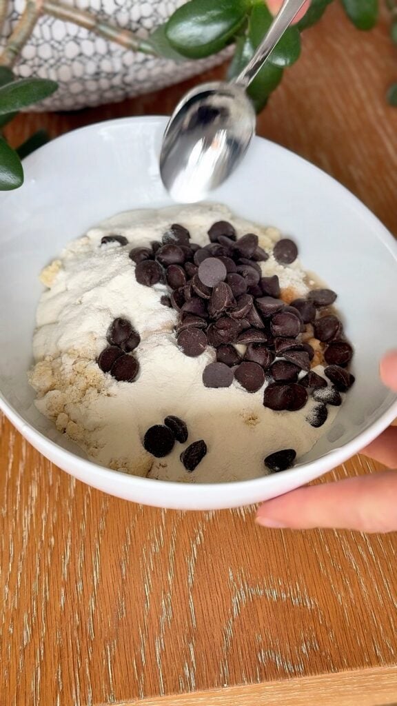 Bowl filled with ingredients for high-protein no-bake cookie dough including cottage cheese, almond flour, protein powder, vanilla, and chocolate chunks.