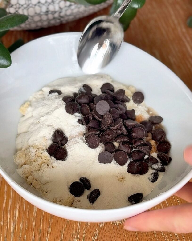 Bowl filled with ingredients for high-protein no-bake cookie dough including cottage cheese, almond flour, protein powder, vanilla, and chocolate chunks.