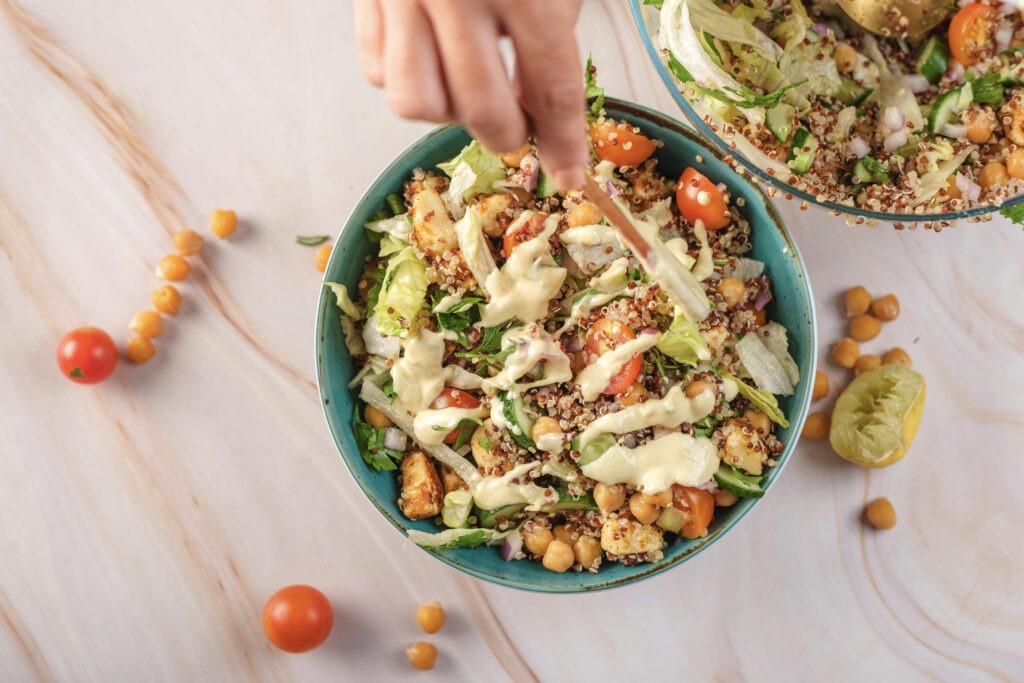 Hand pouring the creamy lemon-tahini dressing over the mixed quinoa and chickpea salad ingredients in a large bowl.