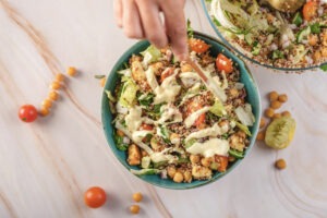 Hand pouring the creamy lemon-tahini dressing over the mixed quinoa and chickpea salad ingredients in a large bowl.