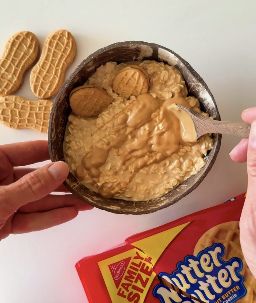 Adding a large swirl of creamy peanut butter into a dark bowl of overnight oats with Nutter Butter cookies on the side.