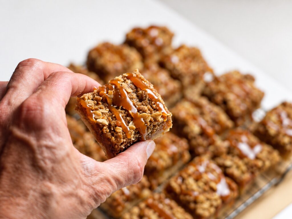 Hand holding a gluten-free apple pie bar topped with oat streusel and salted caramel.
