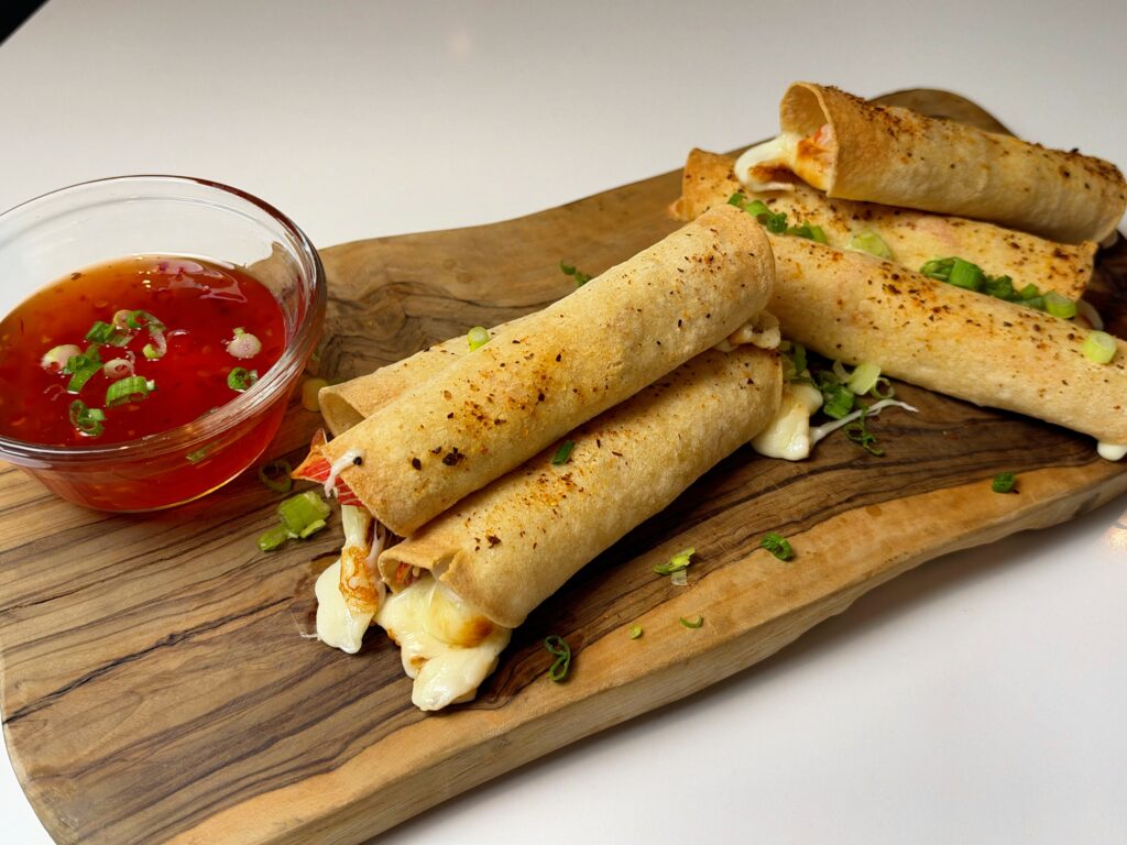 Crispy air fryer crab rangoon mozzarella taquitos arranged on a wooden board with sweet chili sauce.