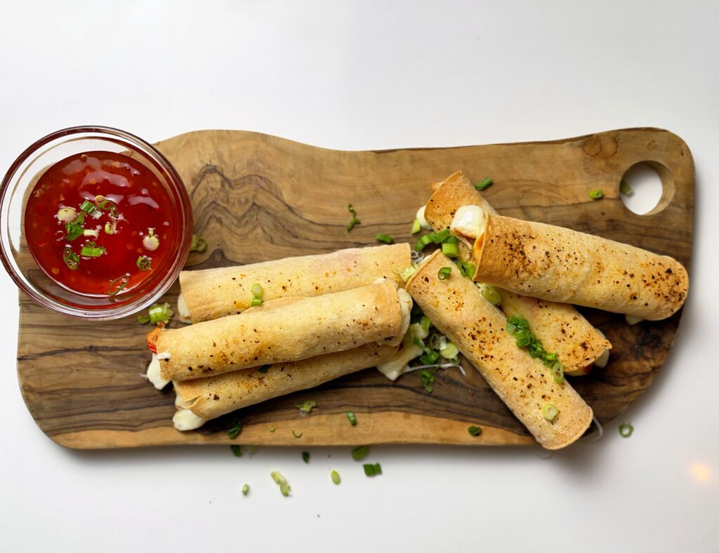 Overhead view of crispy crab rangoon mozzarella taquitos on a wooden board with sweet chili dipping sauce.