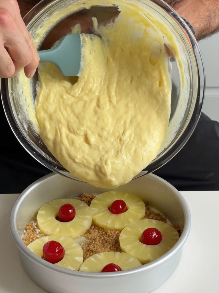 Pineapple protein cake batter being poured over pineapple rings, cherries, and brown sugar in an 8-inch cake pan.
