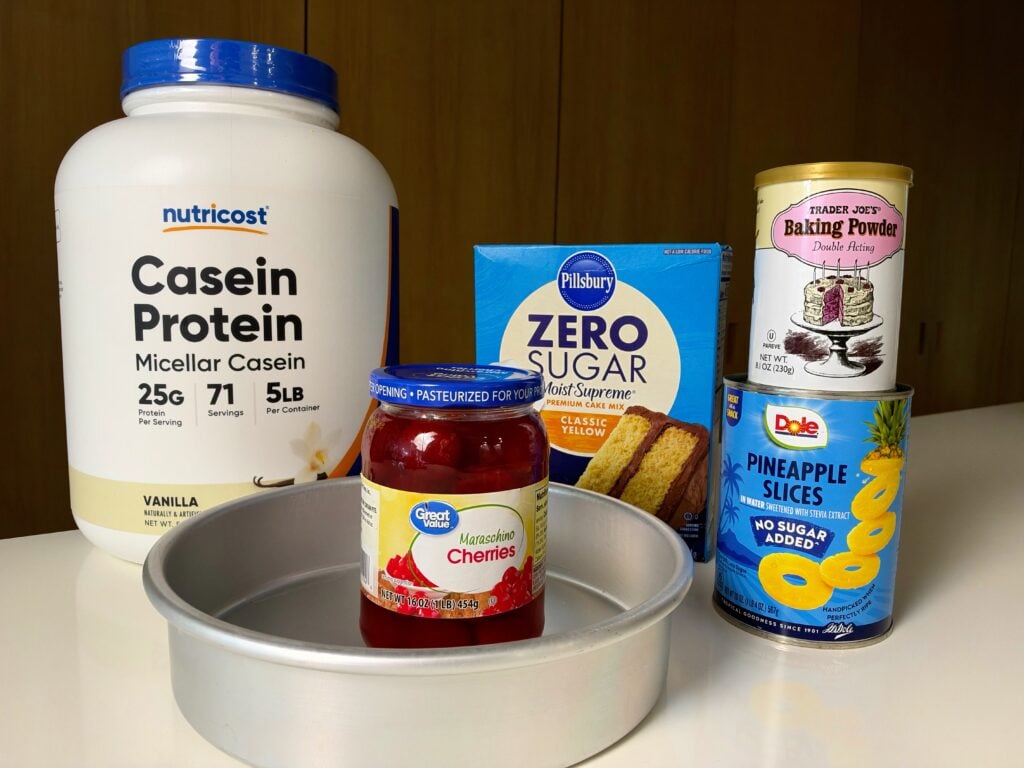 Ingredients for high-protein pineapple upside-down cake including casein protein, zero-sugar cake mix, pineapple rings, cherries, baking powder, and an 8-inch pan.