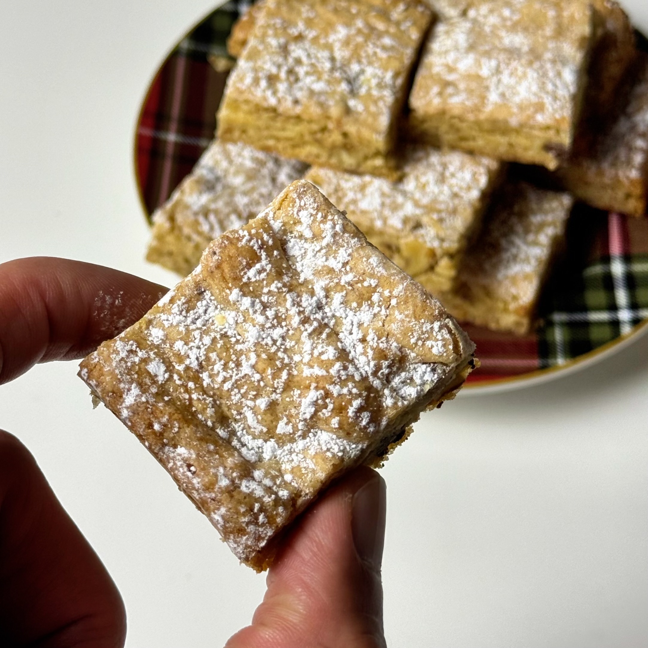Hand holding a powdered sugar-dusted chewy bar with more bars on a plaid plate in the background.