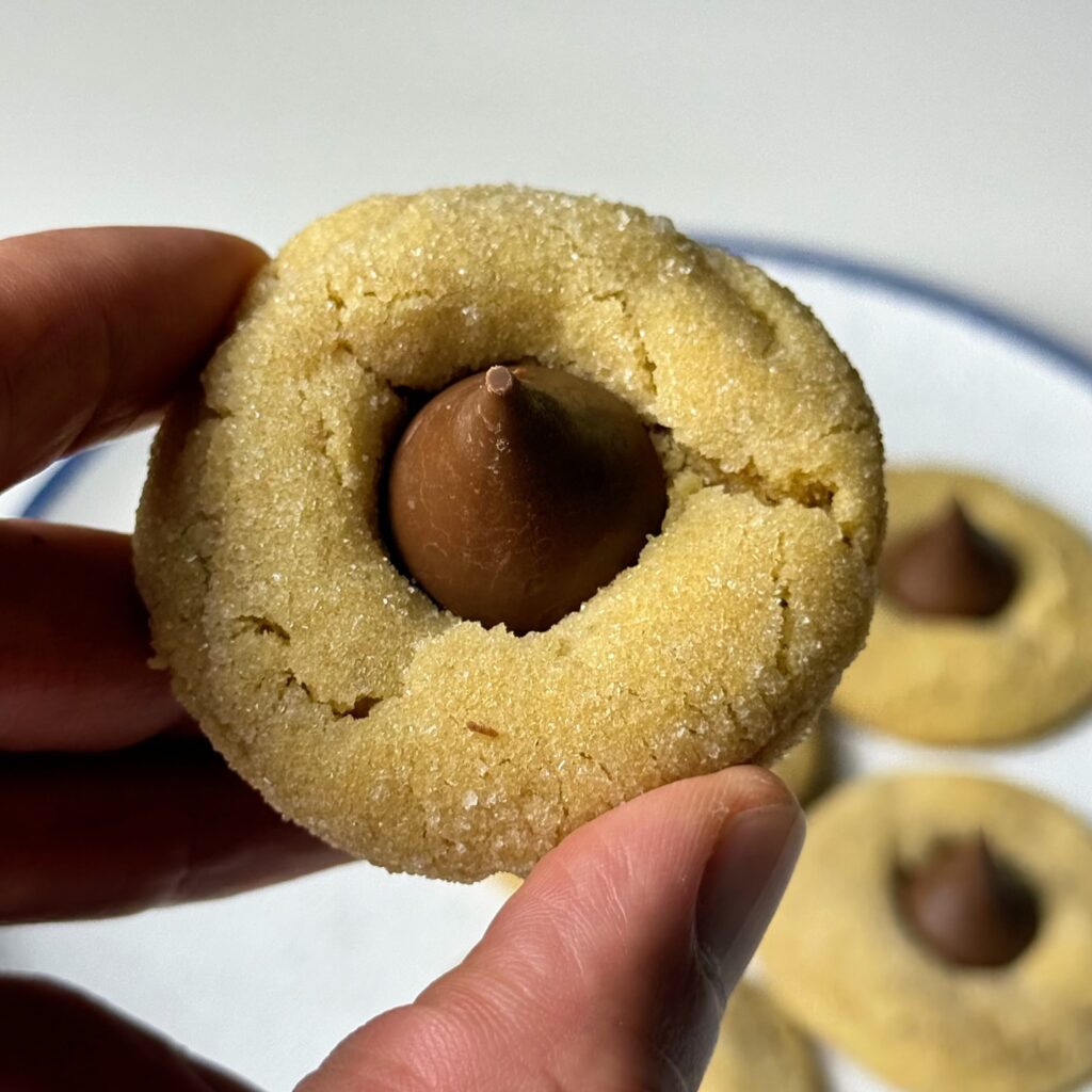 Close-up of a sugar-coated peanut butter blossom cookie held between two fingers with more cookies in the background.