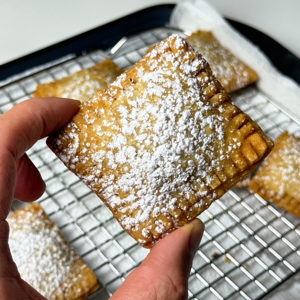 Hand holding a square fried Cuzadache pastry dusted generously with powdered sugar.