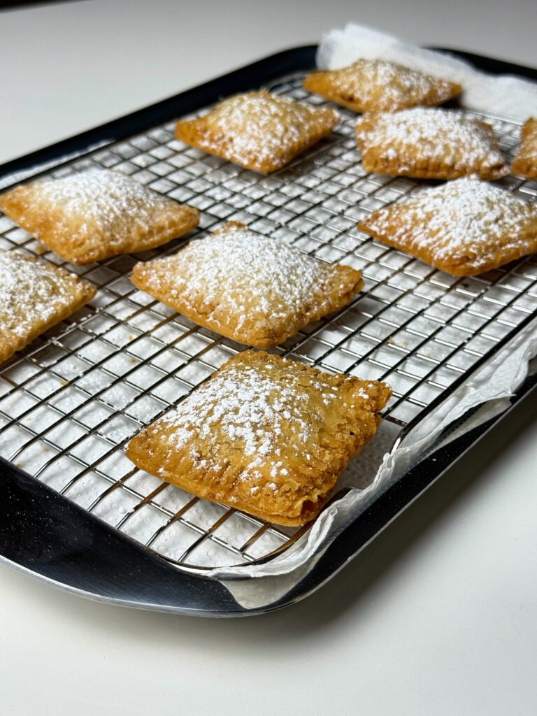 Powdered sugar-dusted Cuzadache pastries cooling on a wire rack over a paper towel-lined tray.