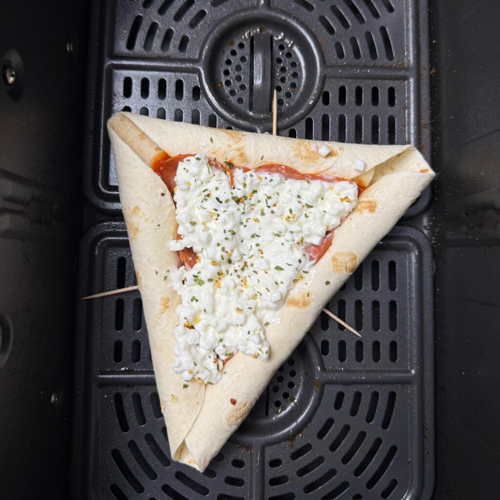Assembled triangular tortilla pizza with folded cheese-stuffed edges, cottage cheese, pepperoni, and herbs in an air fryer basket.