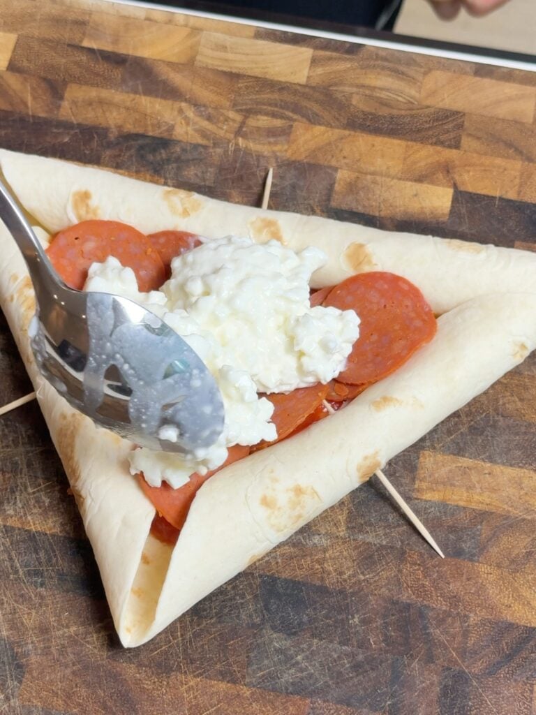Spreading cottage cheese over pepperoni inside a triangular tortilla pizza with folded edges secured by toothpicks.