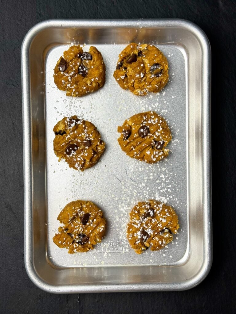 Overhead view of six pumpkin spice protein cookies with fork marks and chocolate chips on a baking sheet.