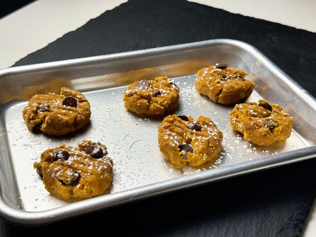 Six pumpkin spice protein cookies with chocolate chips and crosshatch fork marks on a baking sheet.