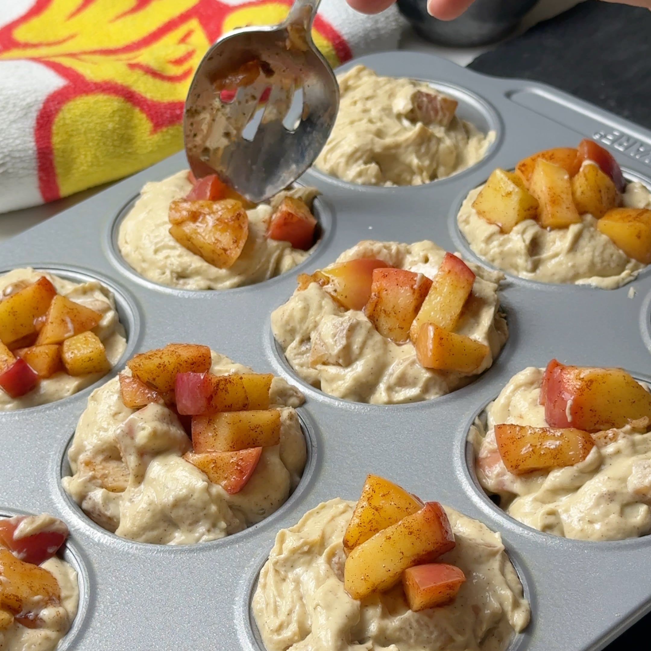 Spoon adding cinnamon topping over diced apples and muffin batter in a lined muffin tin.