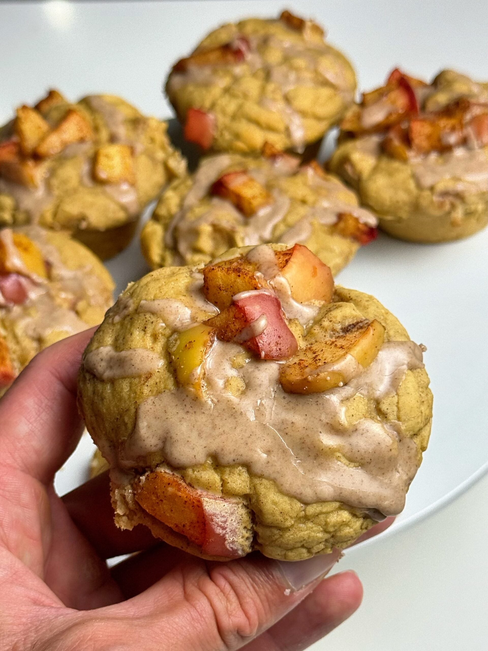 Hand holding an apple pie protein muffin topped with diced apples and cinnamon glaze, with more muffins behind it.