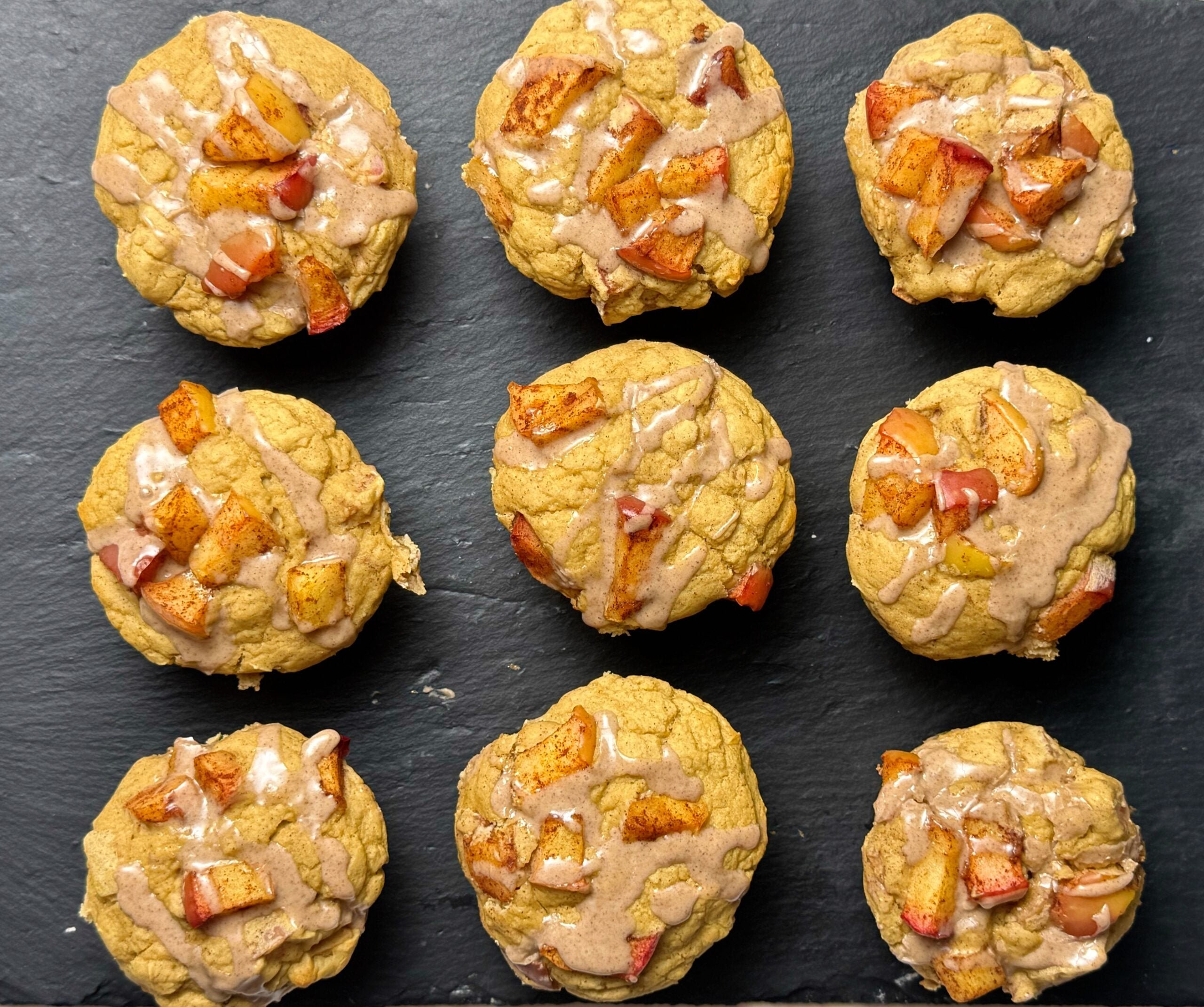 Overhead view of apple pie protein muffins arranged on a dark baking tray with diced apples and glaze on top.