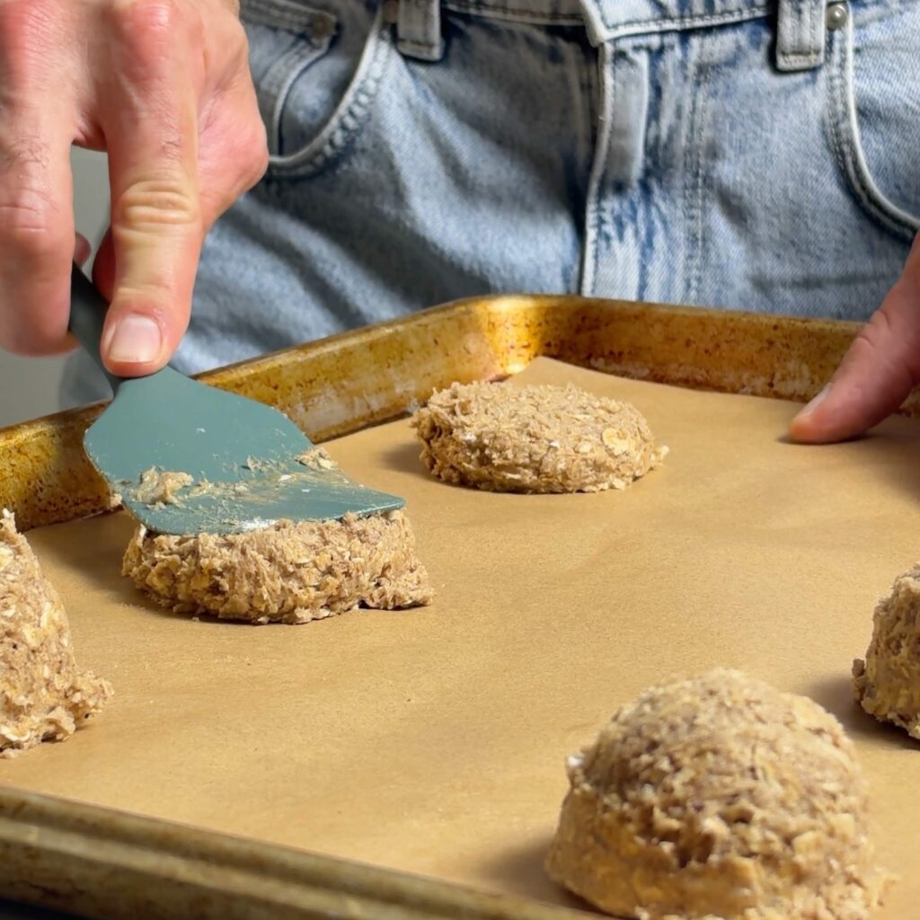 Apple butter oatmeal cookie dough being gently flattened with a silicone spatula on a parchment-lined baking sheet.