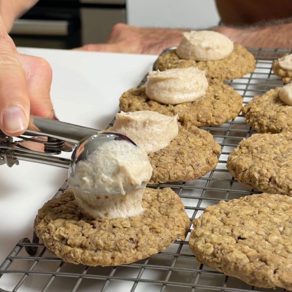 Apple butter cinnamon frosting being spooned onto cooled oatmeal cookies on a wire rack.