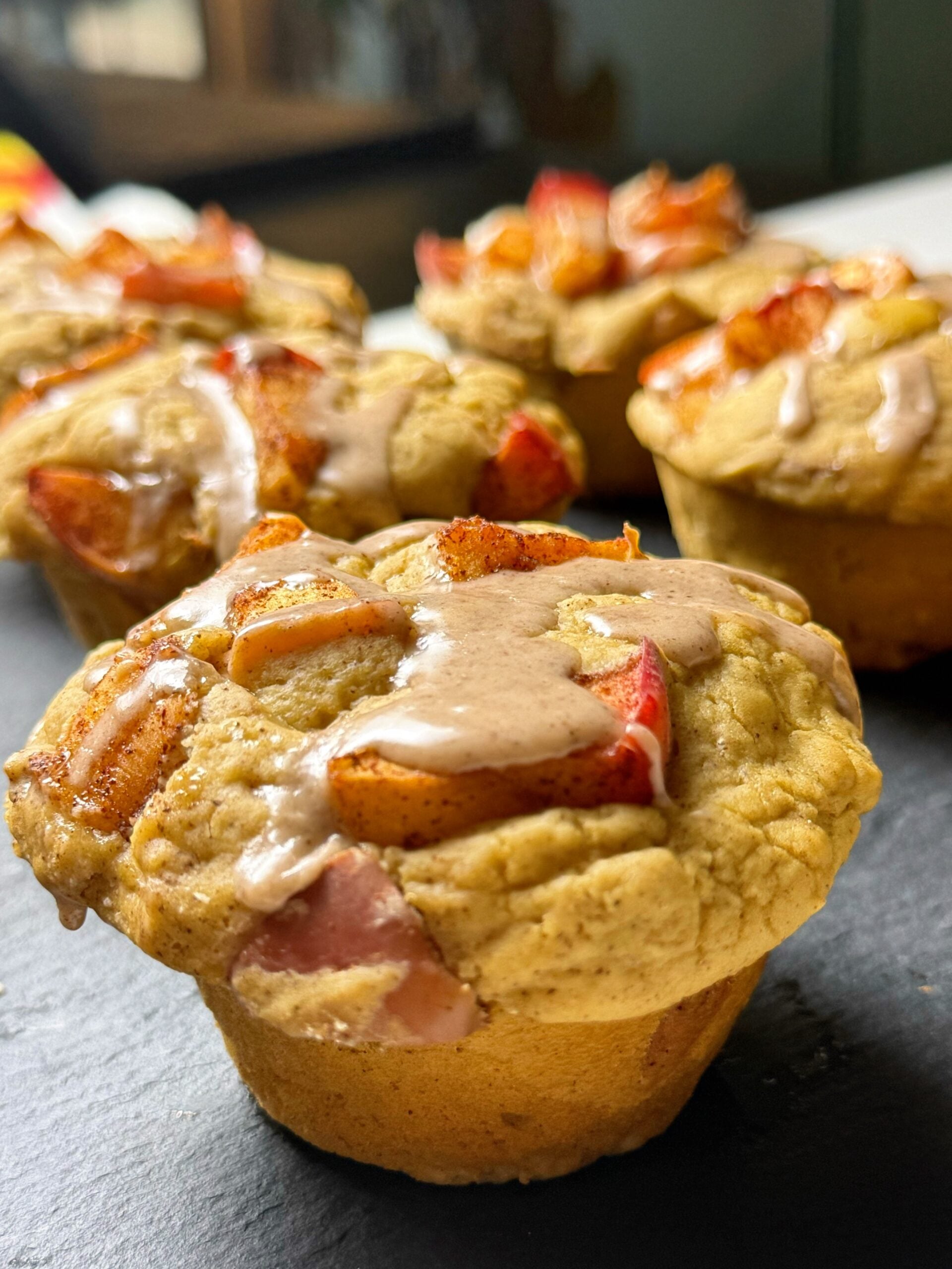 Apple pie protein muffins in paper liners, topped with diced apples and cinnamon glaze.