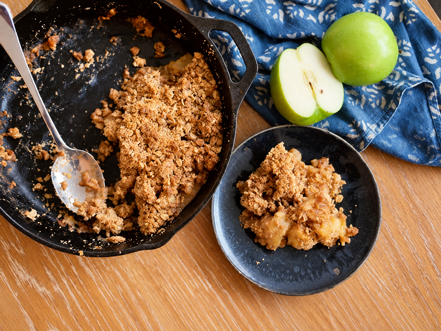 Overhead view of gluten-free apple crisp served from a cast iron skillet onto a dark plate with green apples nearby.