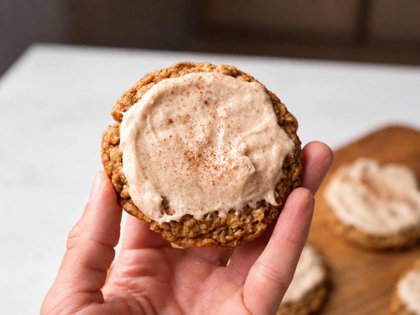 Hand holding a frosted apple butter oatmeal cookie with a tray of cookies in the background.