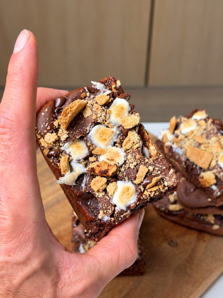 Hand holding a chocolate banana s'mores brownie square with toasted marshmallows, chocolate chips, and graham crackers