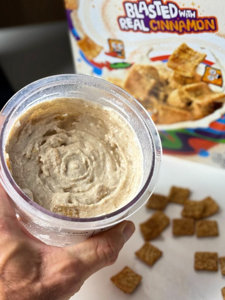 Hand holding a pint of high-protein Cinnamon Toast Crunch Ninja Creami with cereal pieces and the box in the background.