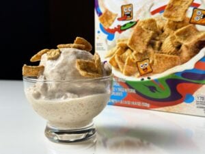High-protein Cinnamon Toast Crunch Ninja Creami topped with cereal pieces in a glass bowl beside the cereal box.