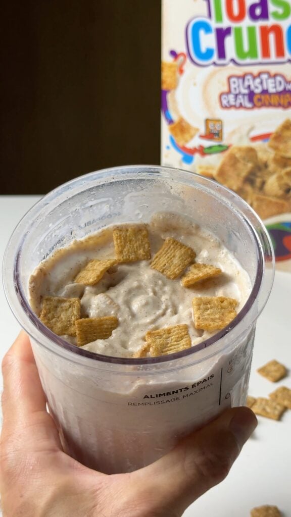Hand holding a Ninja Creami pint filled with high-protein cinnamon ice cream and Cinnamon Toast Crunch cereal pieces.