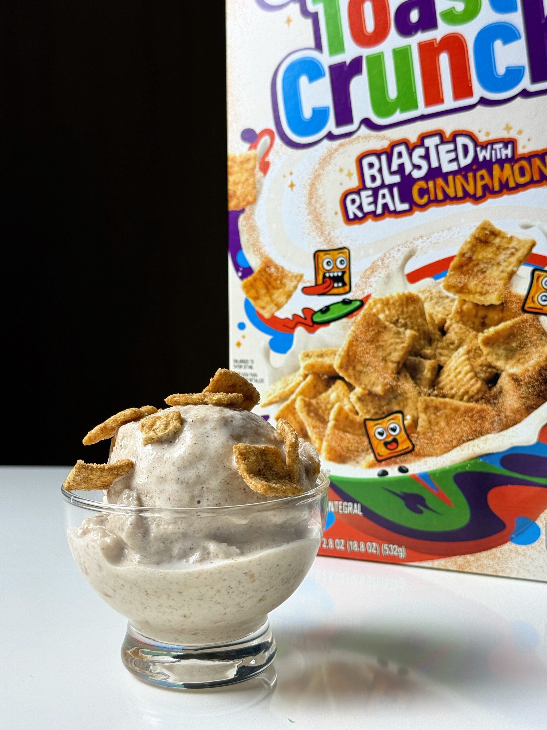 Glass bowl of high-protein Cinnamon Toast Crunch Ninja Creami topped with cereal pieces in front of the cereal box.