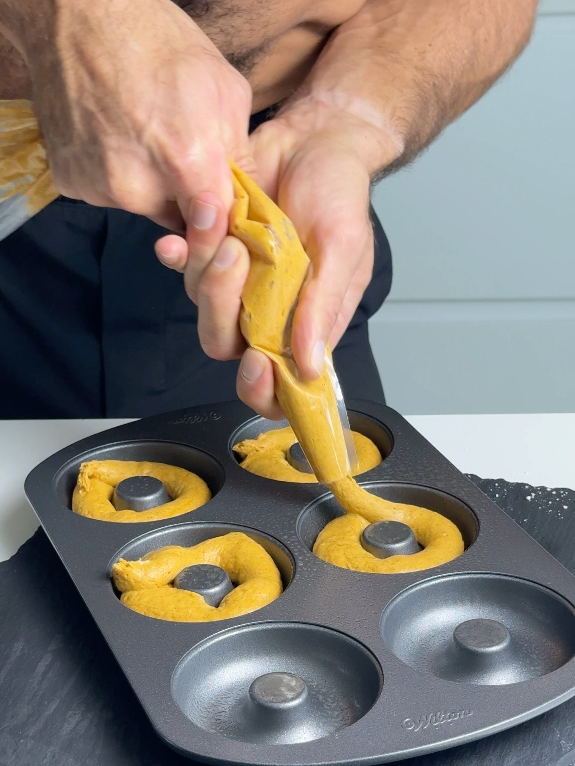 Hands piping thick pumpkin spice donut batter into a donut pan
