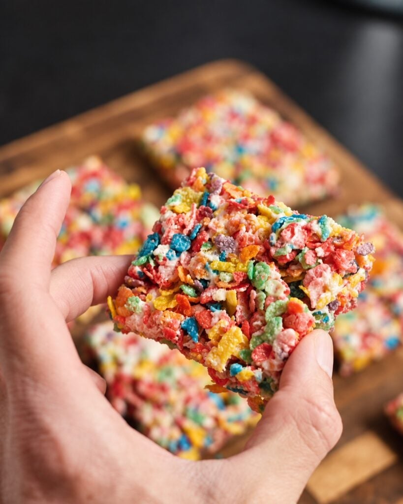 Two hands holding one thick protein Fruity Pebbles rice crispy treat with more bars behind it.