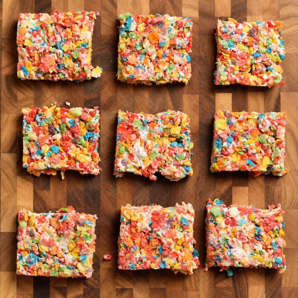 Nine high-protein Fruity Pebbles rice crispy treats arranged on a wooden cutting board.