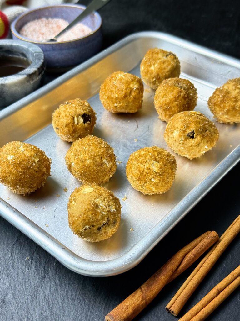 Pumpkin spice protein balls with visible oats and chocolate chips arranged on a metal tray beside cinnamon sticks.