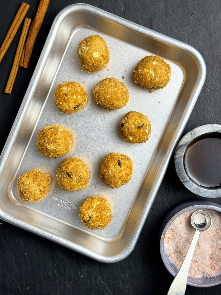 Overhead view of ten pumpkin spice protein balls on a metal tray, with cinnamon sticks and small bowls alongside.