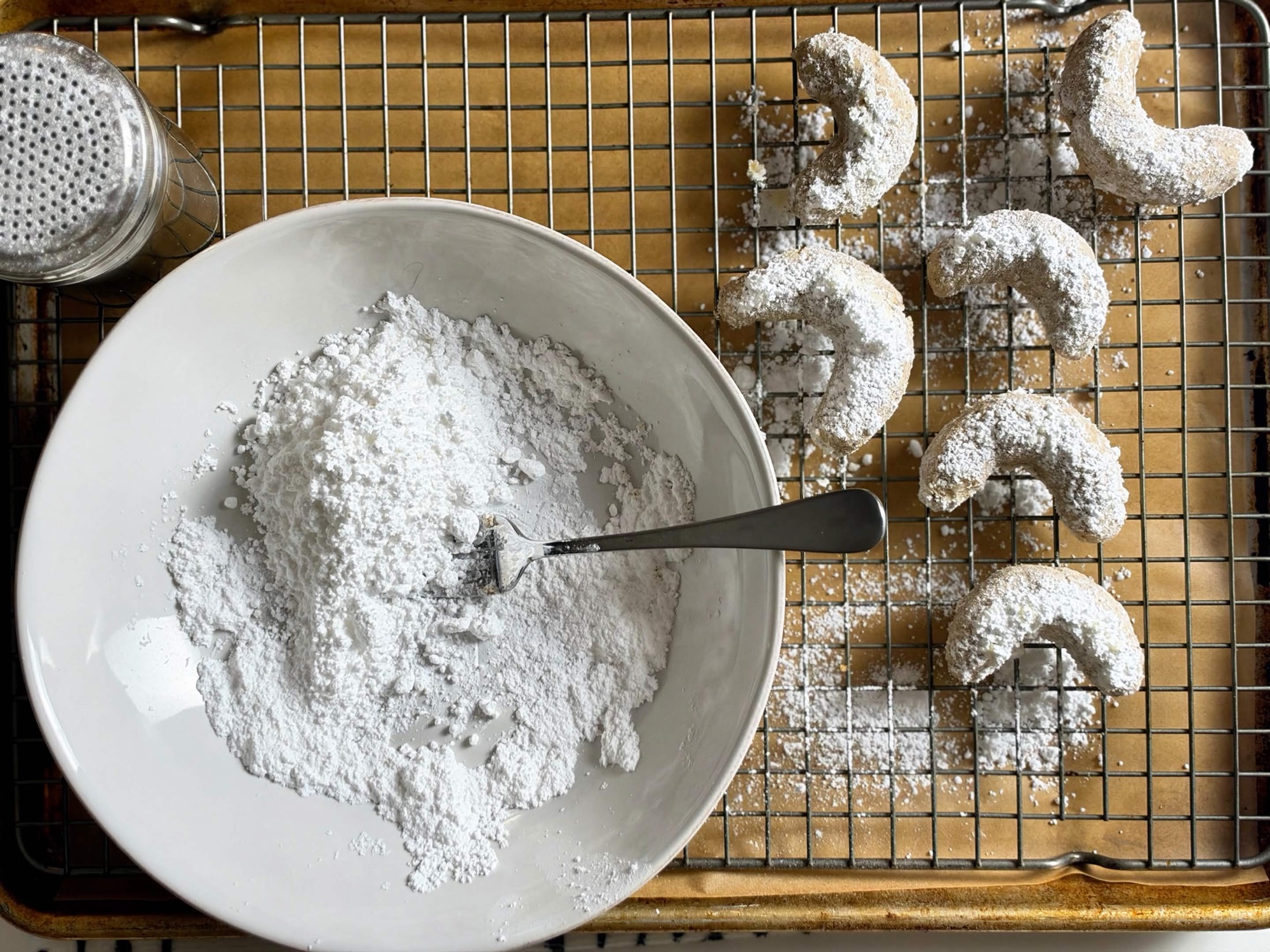 Pecan crescent cookies being coated in powdered sugar beside a cooling rack
