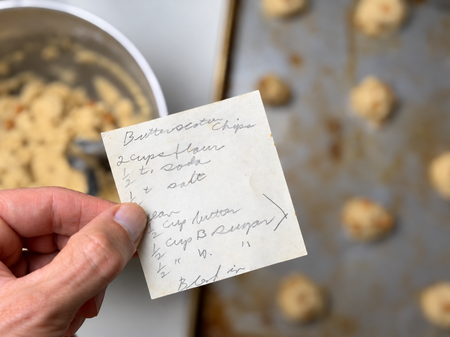 Grandma’s handwritten butterscotch chip cookie recipe card held over cookie dough and a baking sheet