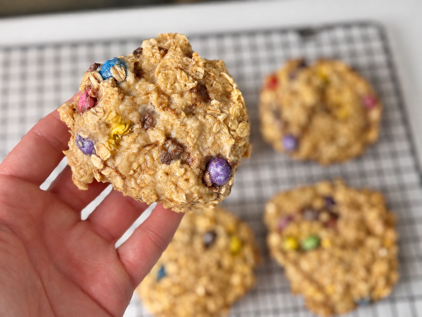 Hand holding a banana oatmeal M&M breakfast cookie over a cooling rack