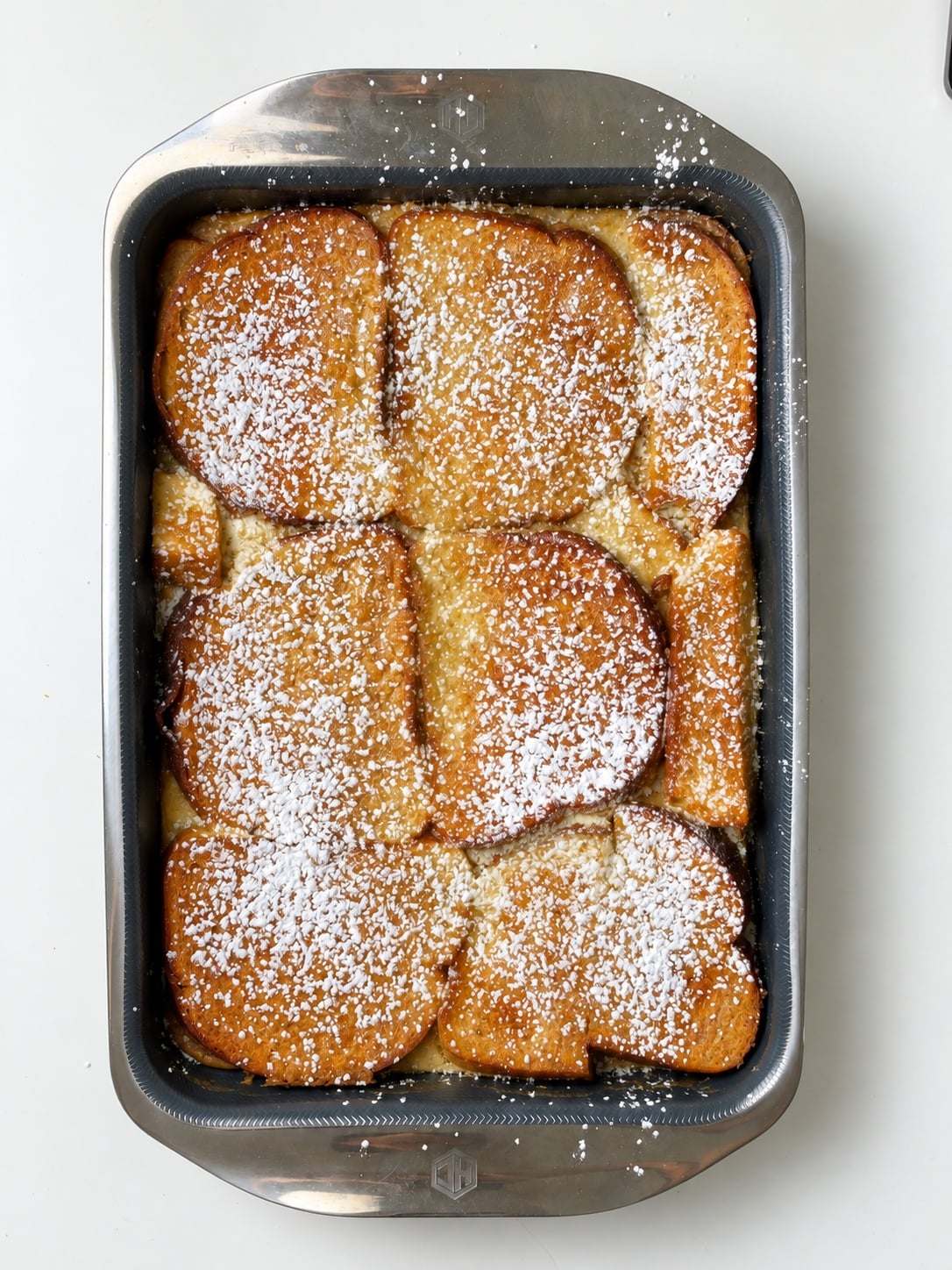 Overhead view of a high-protein French toast casserole dusted with powdered sweetener in a rectangular baking pan.